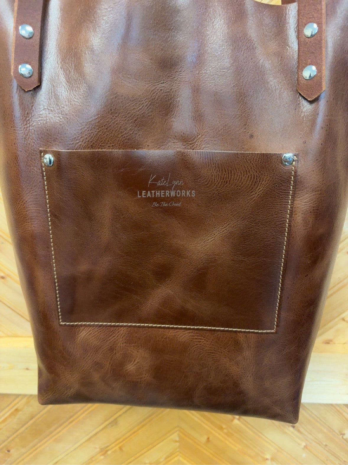 Brown back of Bristow bookbag showing its exterior patch pocket, KateLynn Leatherworks logo and silver-tone rivets.