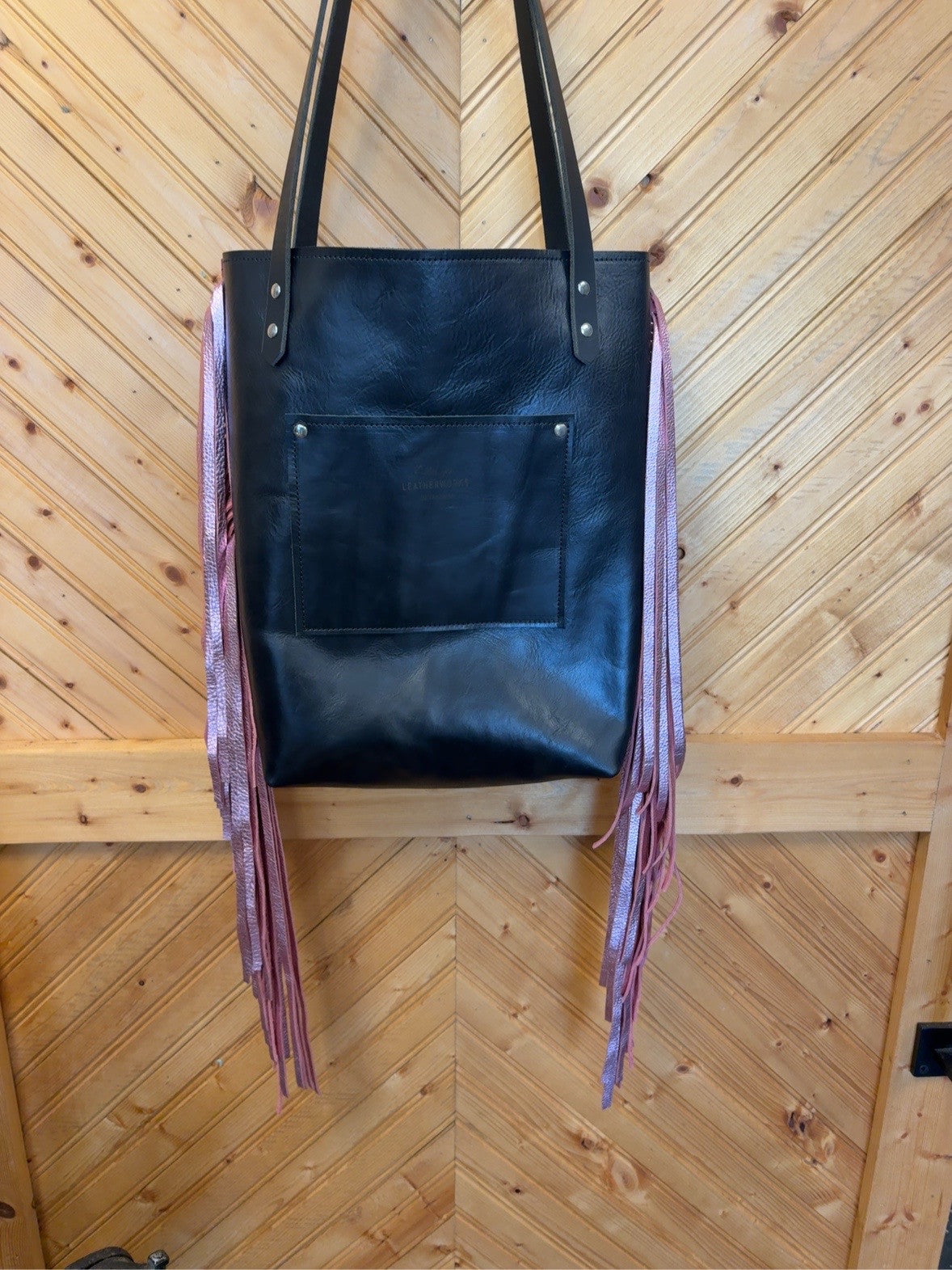 Black back of Bristow bookbag showing exterior patch pocket, long pink side fringe and black shoulder straps.