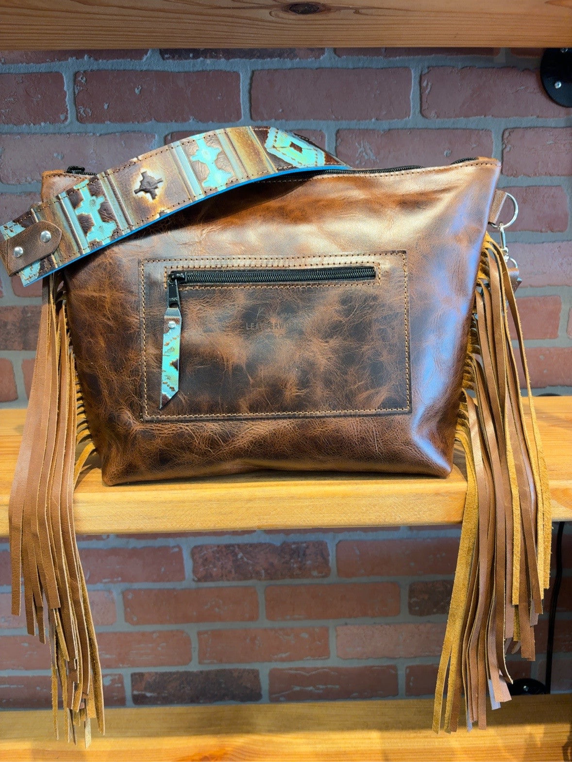 Back of brown KC bag showing rear zipper pocket, patterned strap and side fringe.