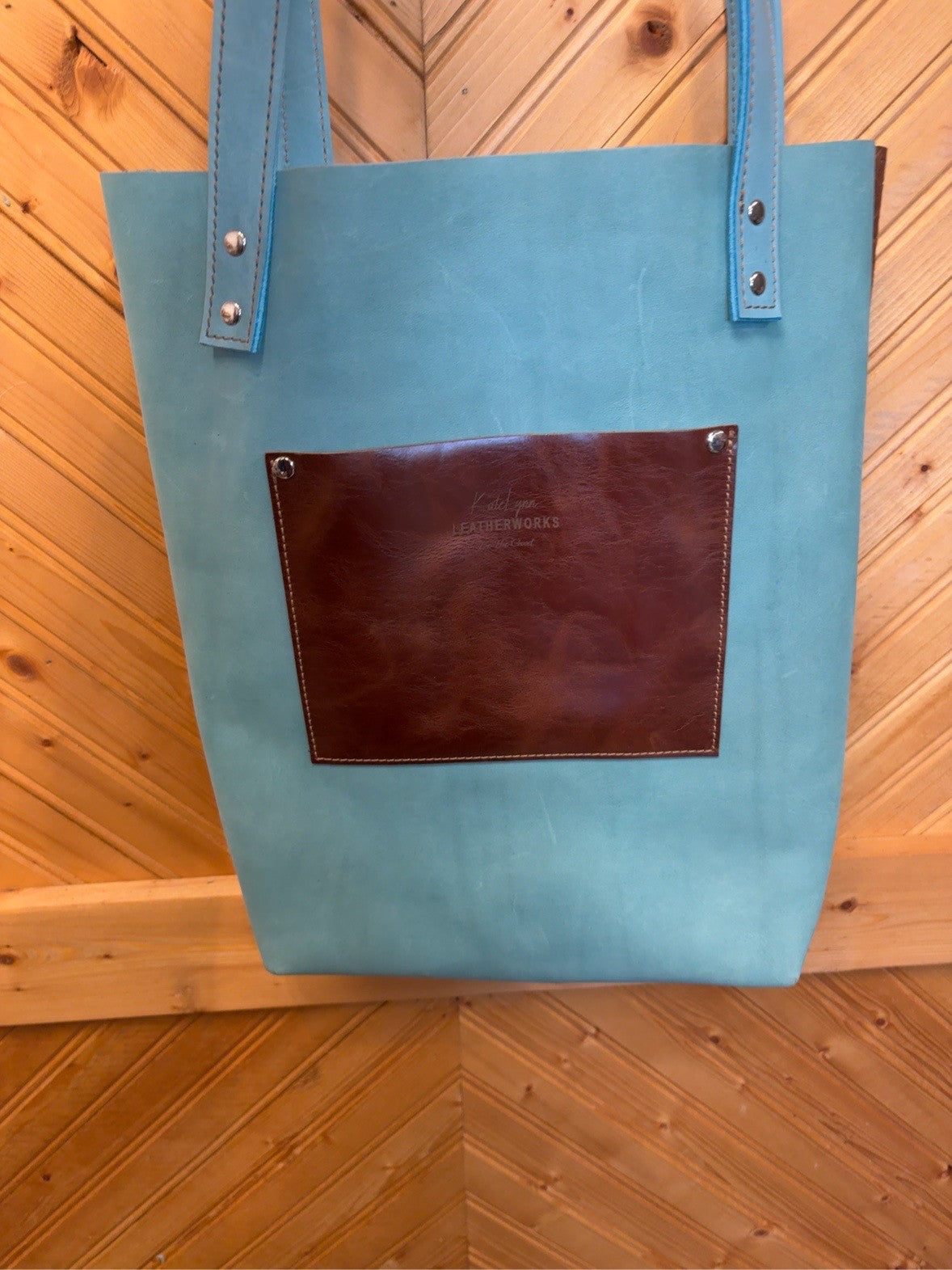 Turquoise back of the Bristow Bookbag with a reddish-brown exterior pocket, KateLynn Leatherworks stamp, turquoise straps and silver-tone rivets.