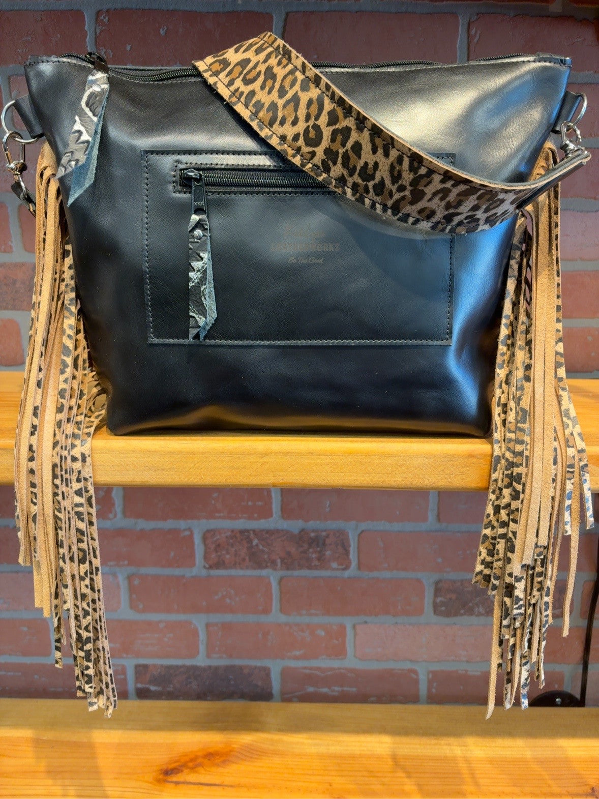 Back of the black KC bag showing the exterior zipper pocket, leopard-patterned shoulder strap and side fringe.