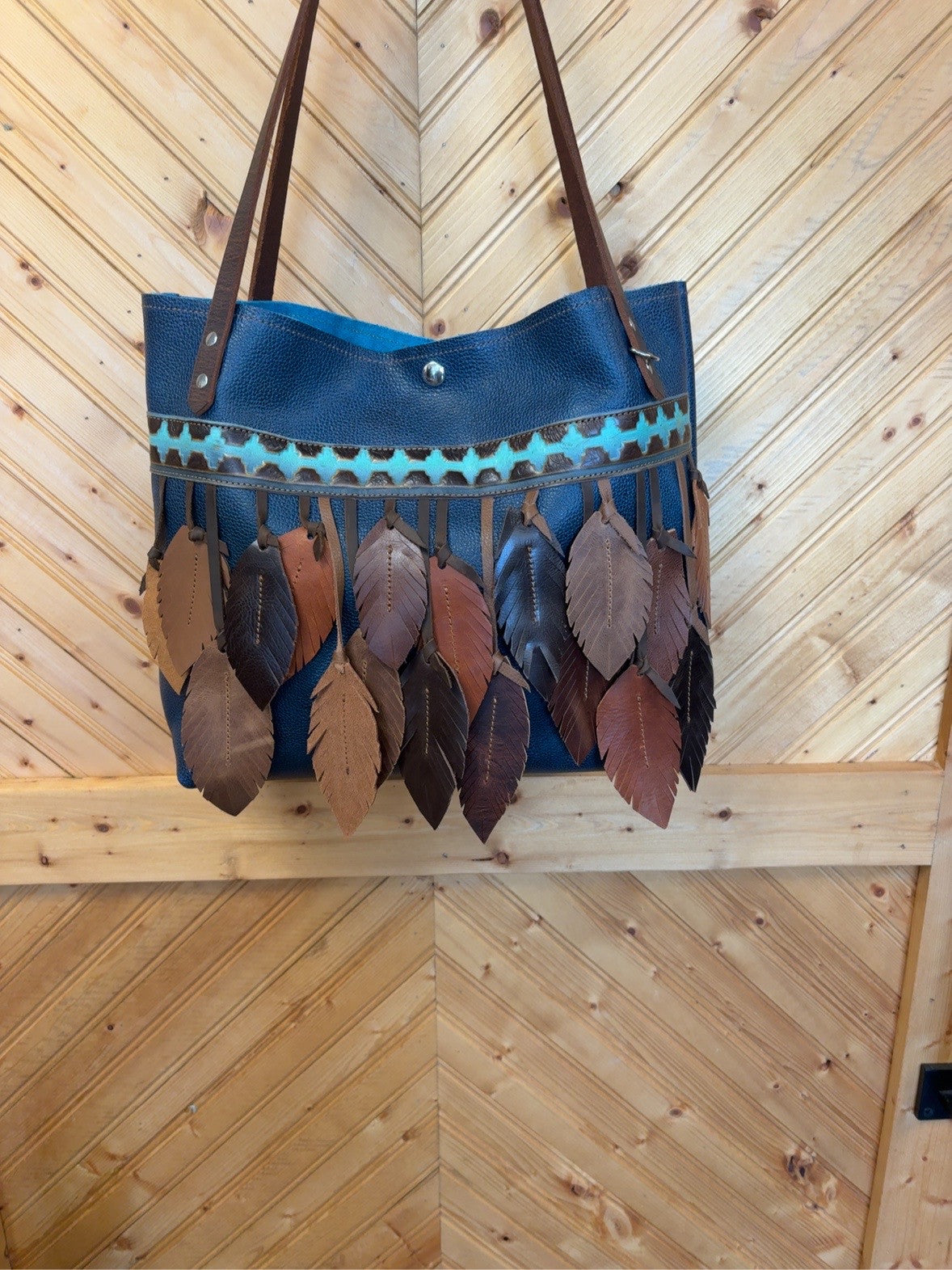 Front of the blue Tucson tote with turquoise-and-brown geometric trim, layered brown leather feathers and brown shoulder straps.
