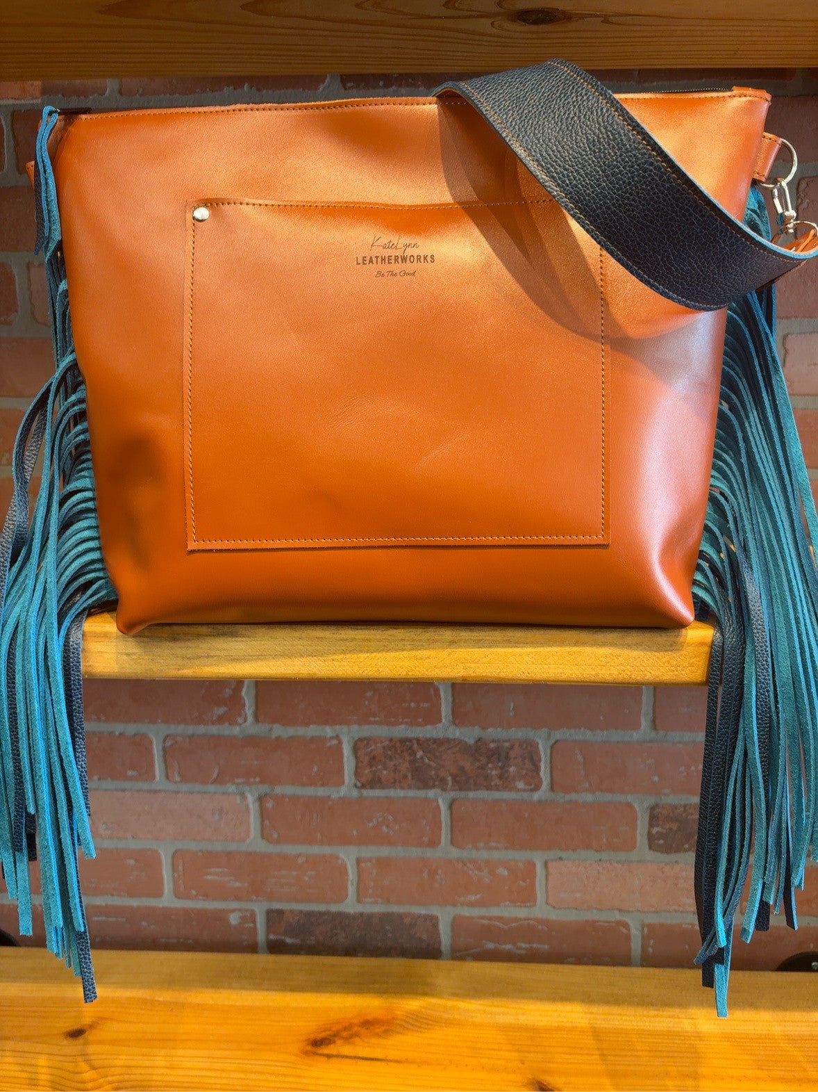 Back of the burnt orange Tucson tote showing the exterior slip pocket, wide blue strap and blue side fringe.