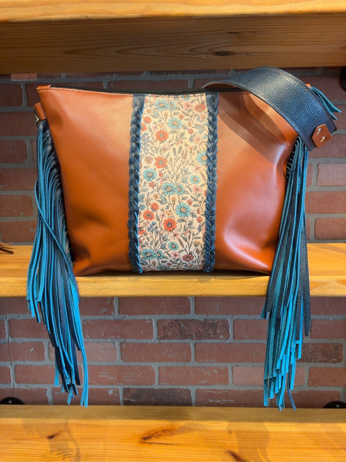 Front of the burnt orange Tucson tote with a floral-pattern center, braided stripes, blue side fringe and a wide blue strap.